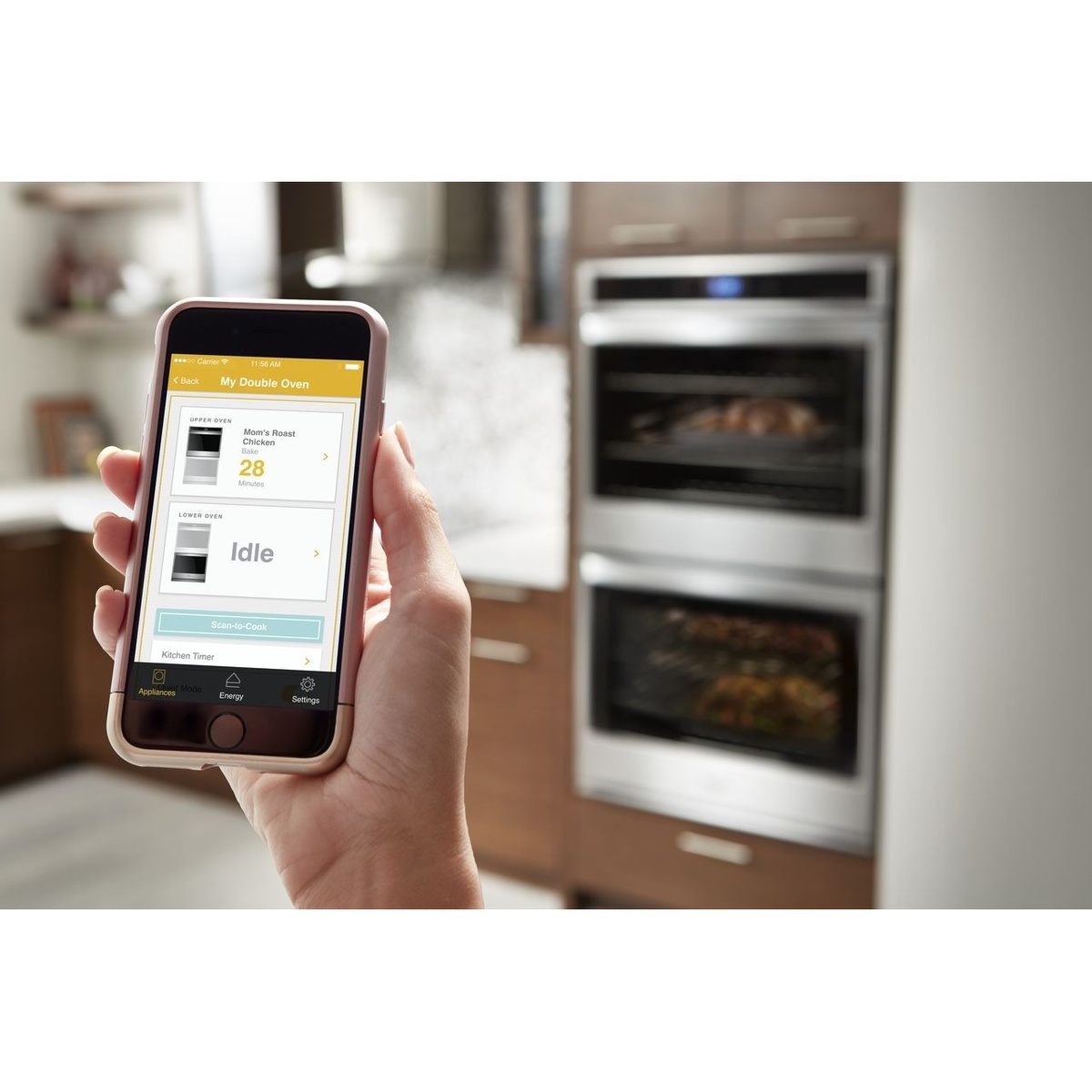 Hand holding a smartphone showing the Whirlpool app screen for a double oven, with a built-in double wall oven in the kitchen background