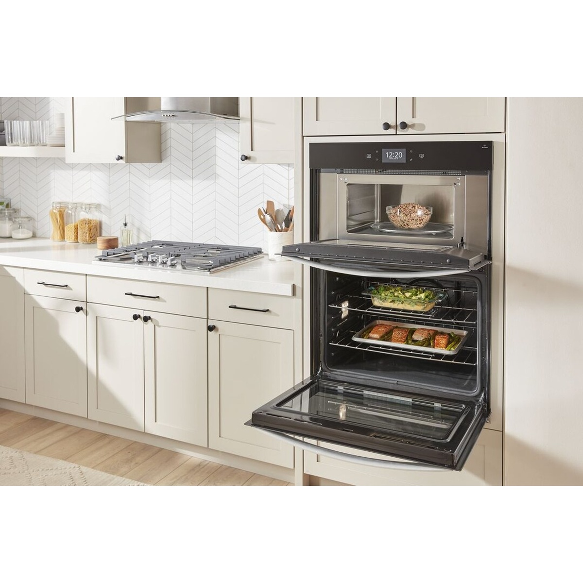 Whirlpool wall oven microwave combo installed in a bright white kitchen with both compartments open, microwave holding a bowl and oven loaded with salmon and vegetables on two racks
