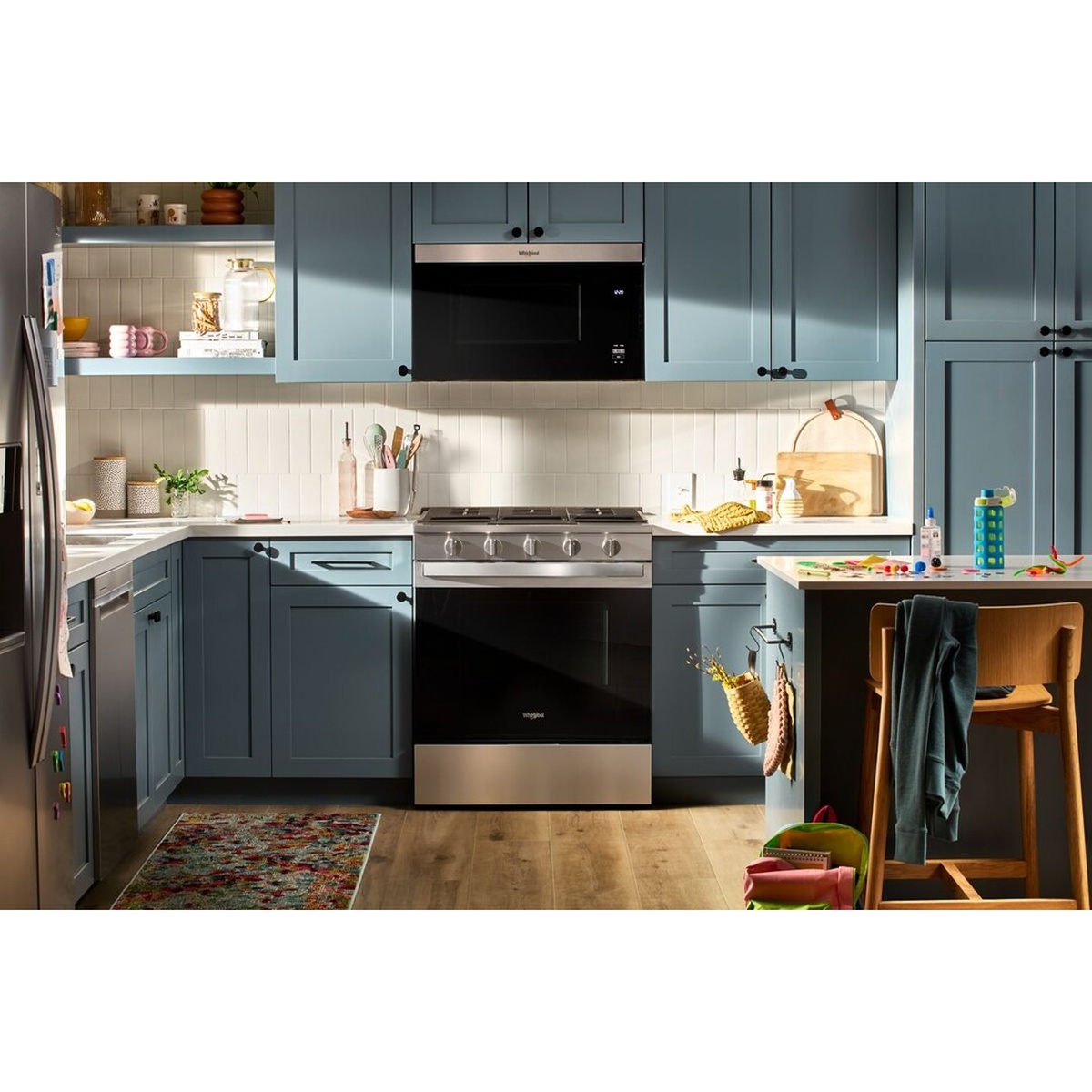 Whirlpool gas slide-in range installed in a lived-in family kitchen with blue cabinetry, an over-the-range microwave, and a busy countertop