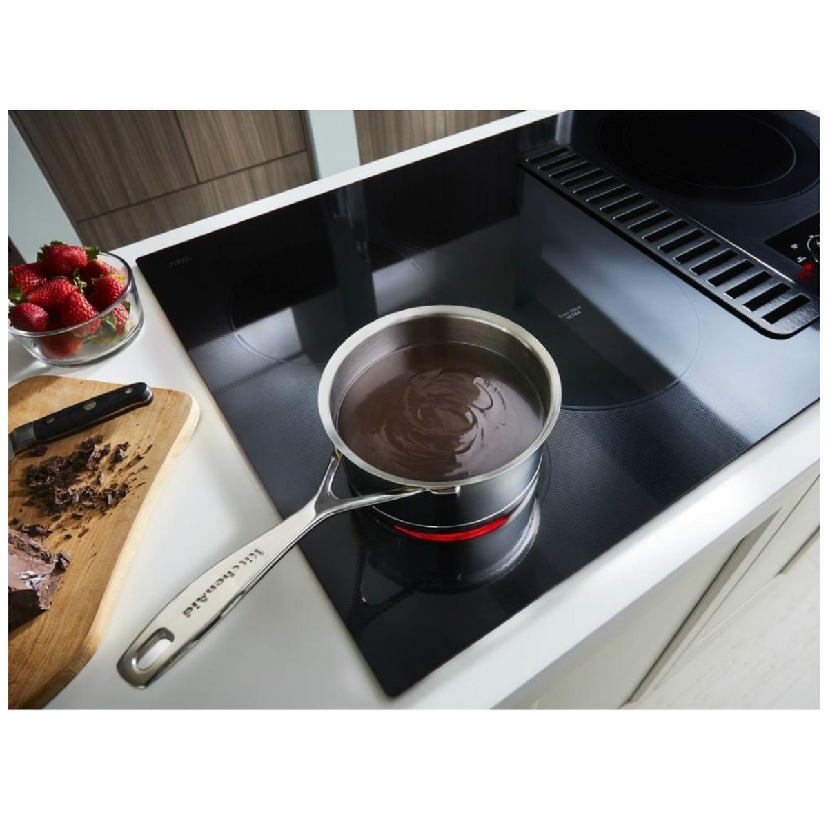 Lifestyle close-up of a KitchenAid saucepan melting chocolate on the Even-Heat Ultra element of the KCED600GBL, installed in a white kitchen counter with the downdraft grate and knobs visible in the background