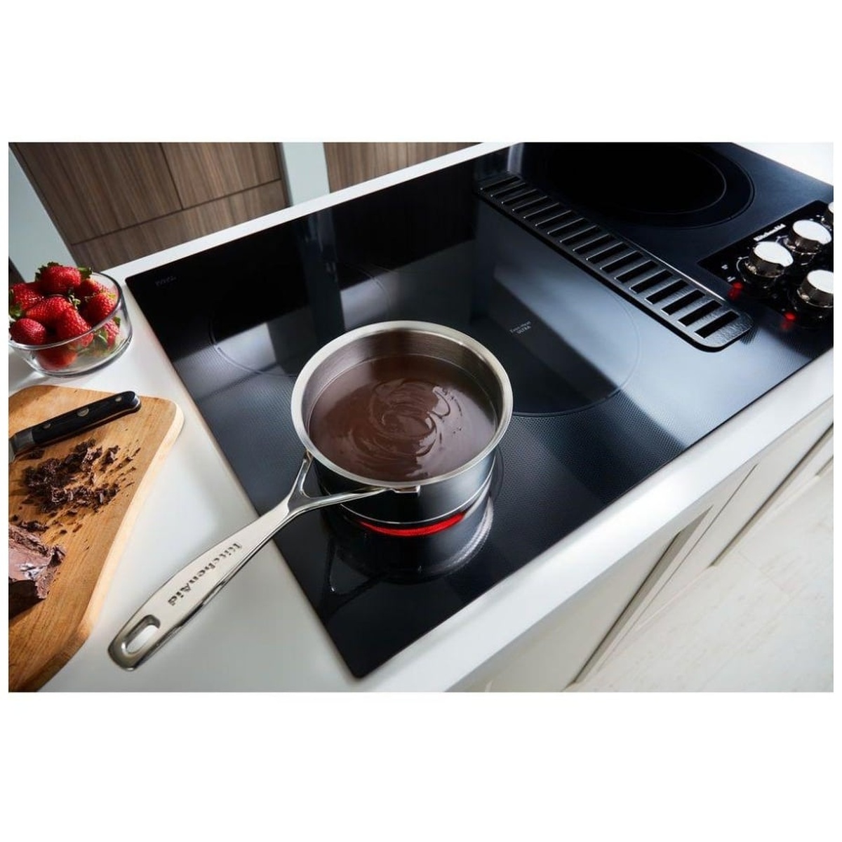 Lifestyle shot of the KitchenAid KCED606GSS installed in a white kitchen with a stainless saucepan of melted chocolate on the cooktop surface, strawberries and a cutting board visible beside it