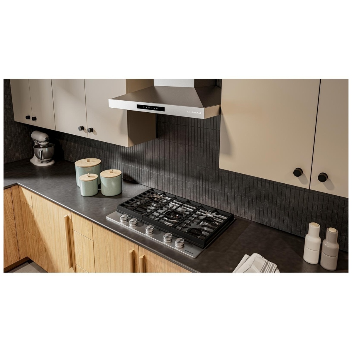 KitchenAid KCGK530SSS gas cooktop installed in a modern kitchen with wood cabinetry, dark countertops, and a KitchenAid range hood above