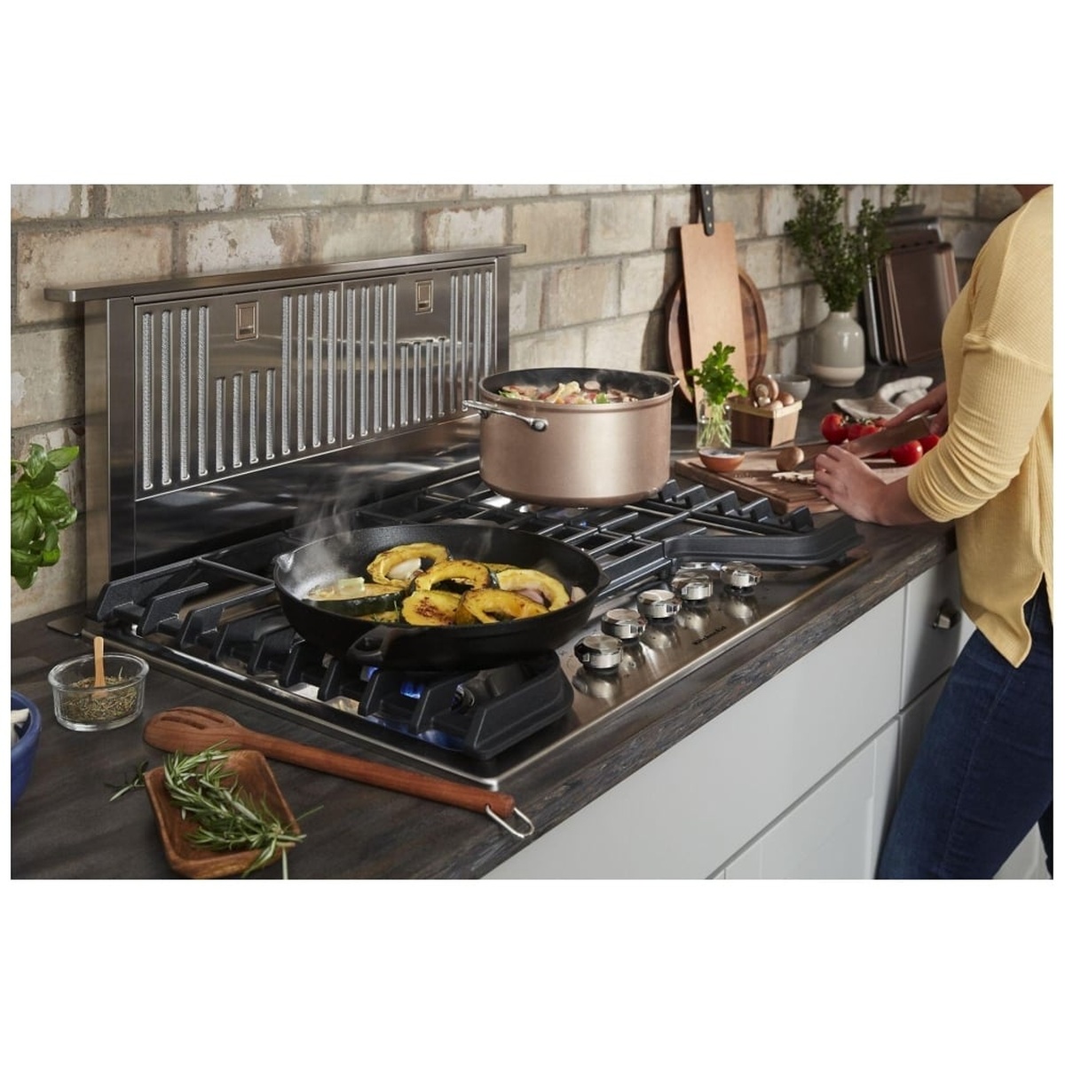 KitchenAid KXD4630YSS retractable downdraft raised behind a gas cooktop, capturing steam and smoke while a person cooks in an open kitchen
