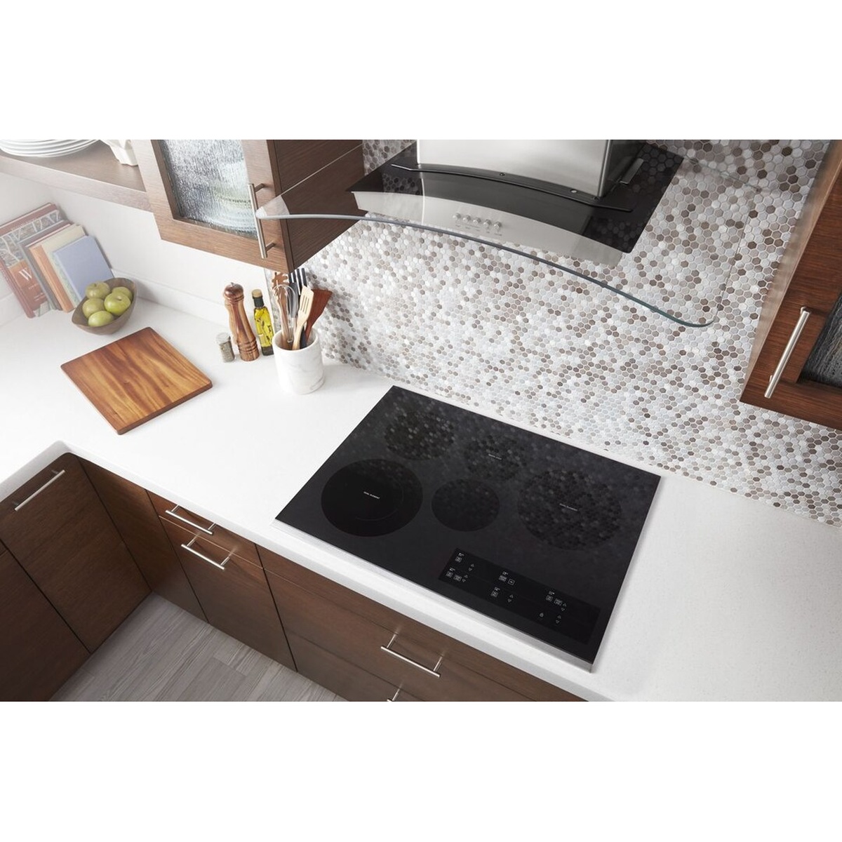 Overhead lifestyle shot of the Whirlpool electric cooktop installed in a modern kitchen with dark wood cabinets, white countertops, mosaic tile backsplash, and a curved range hood above.