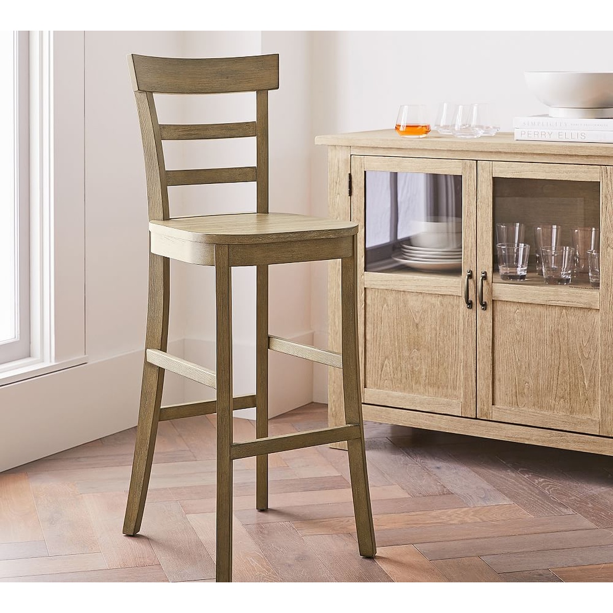 Liam bar stool in Weathered Grey finish styled beside a wood sideboard with glassware in a bright living and dining space