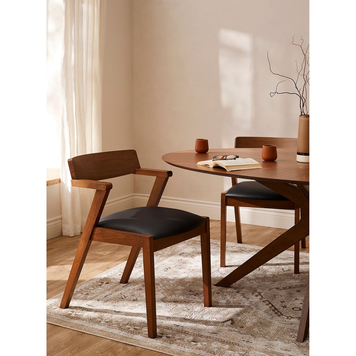 Two Zola dining chairs with walnut oak frames and black vegan leather seats arranged around a round wood dining table in a warm, naturally lit dining room