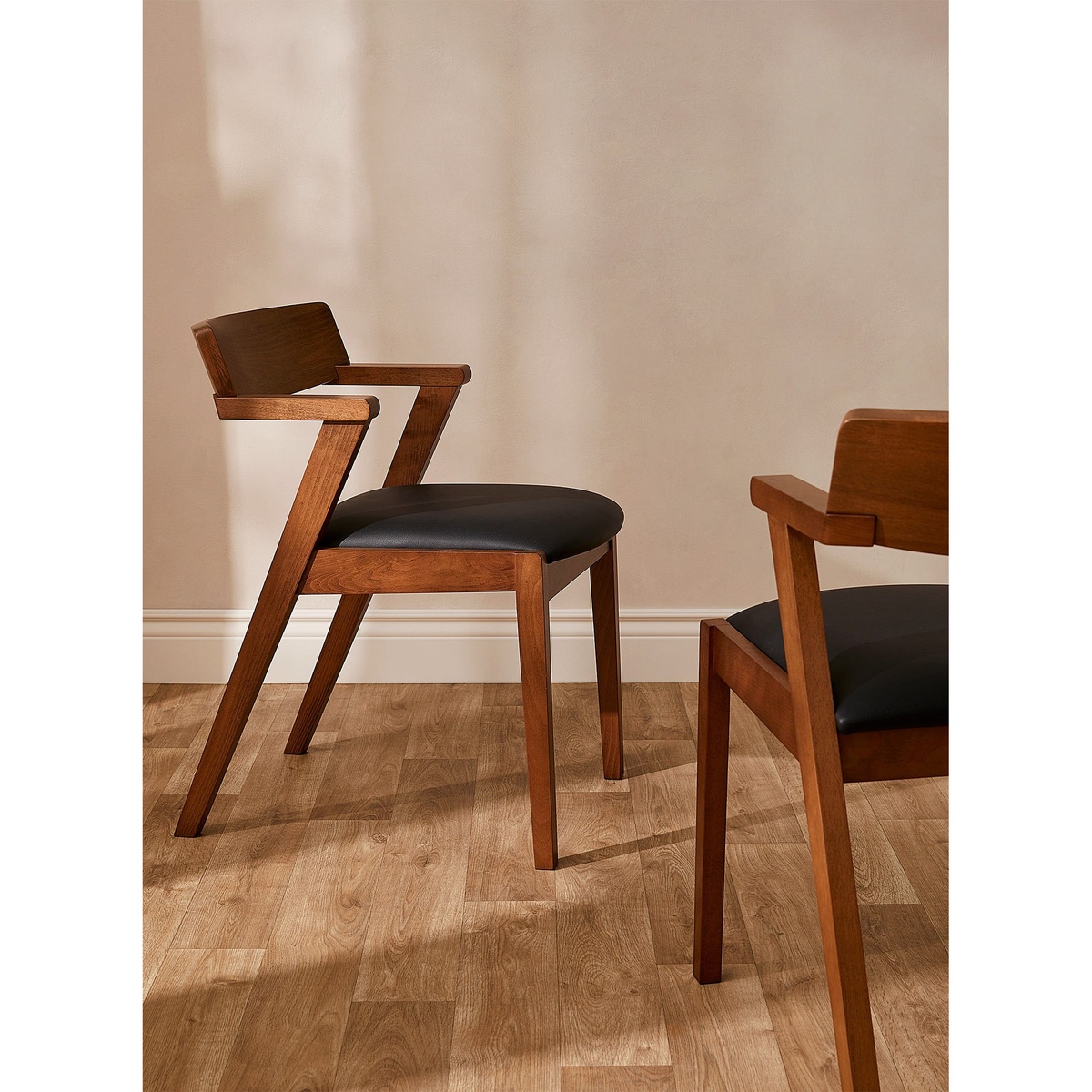 A pair of Zola walnut-and-black dining chairs displayed on a light hardwood floor, showcasing the sculptural frame construction and vegan leather seats from multiple angles