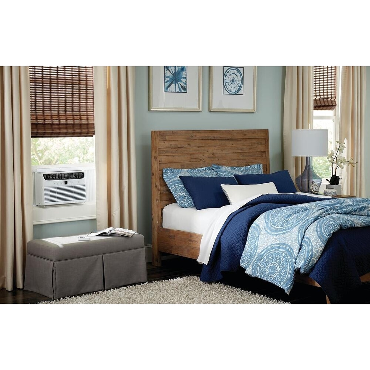Frigidaire FHWW063WB1 window air conditioner installed in a bedroom window alongside a wood-headboard bed with blue bedding, showing the unit in a real living environment