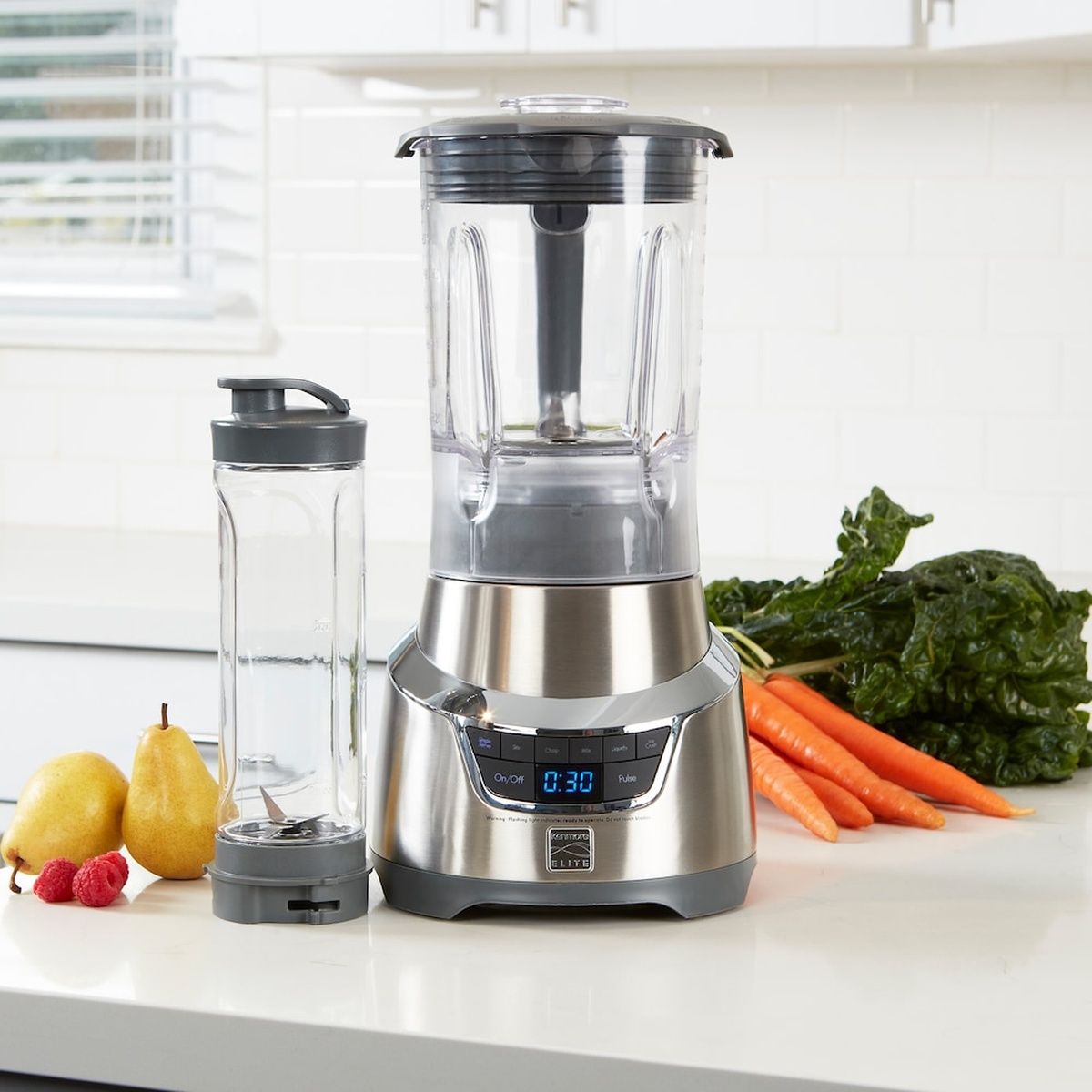 Kenmore Elite countertop blender in stainless finish with large blending jar and a separate single-serve cup on a kitchen counter with fruit and vegetables.