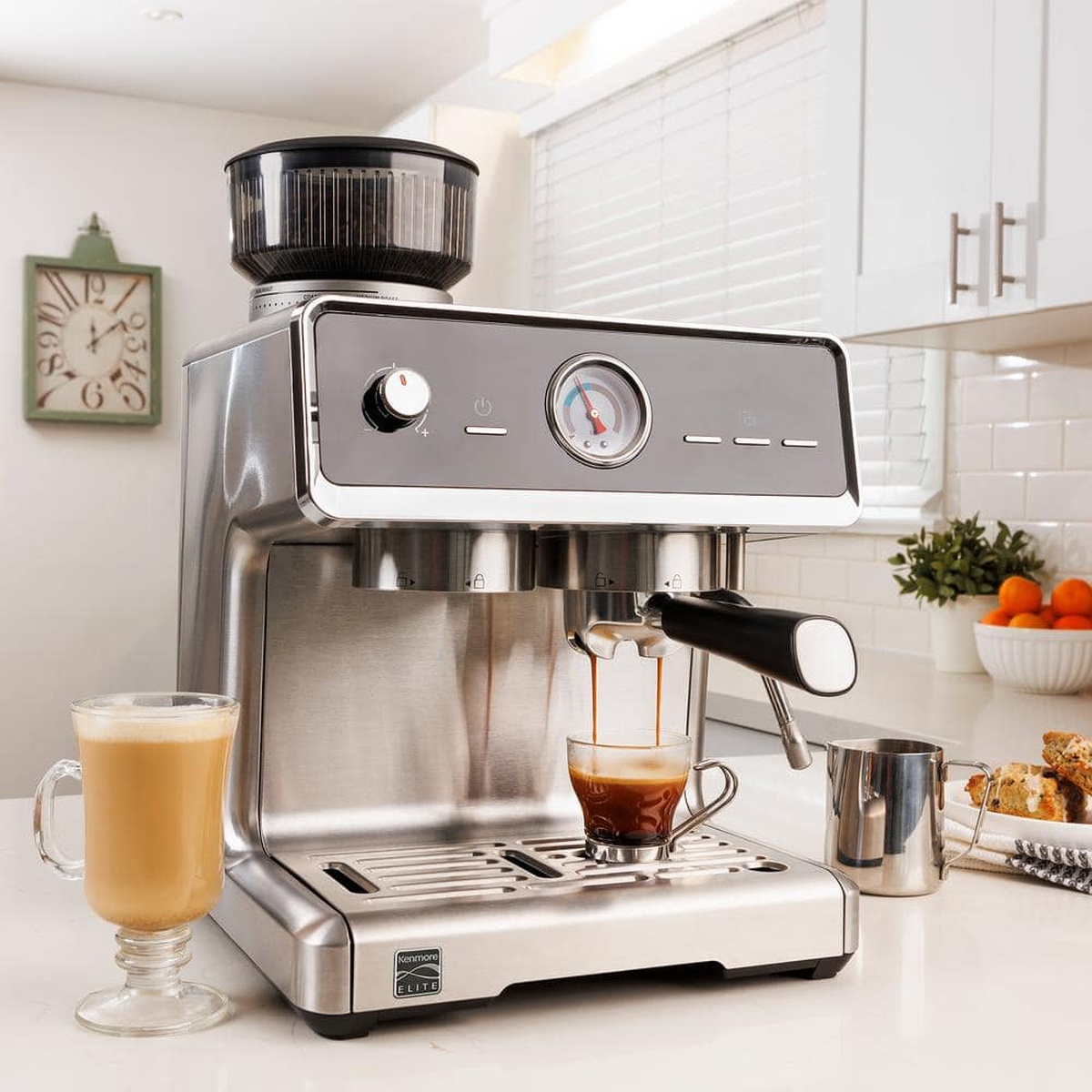 Kenmore Elite espresso machine on a kitchen counter pulling espresso into a glass cup with drinks and a milk pitcher nearby