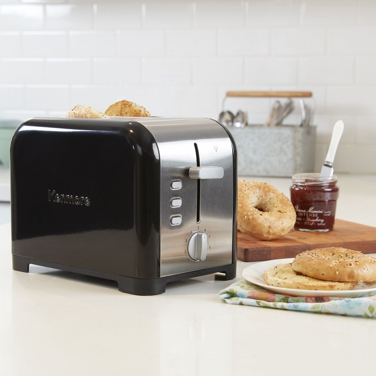 Kenmore black and stainless steel 2-slice toaster on a kitchen counter beside bagels and jam