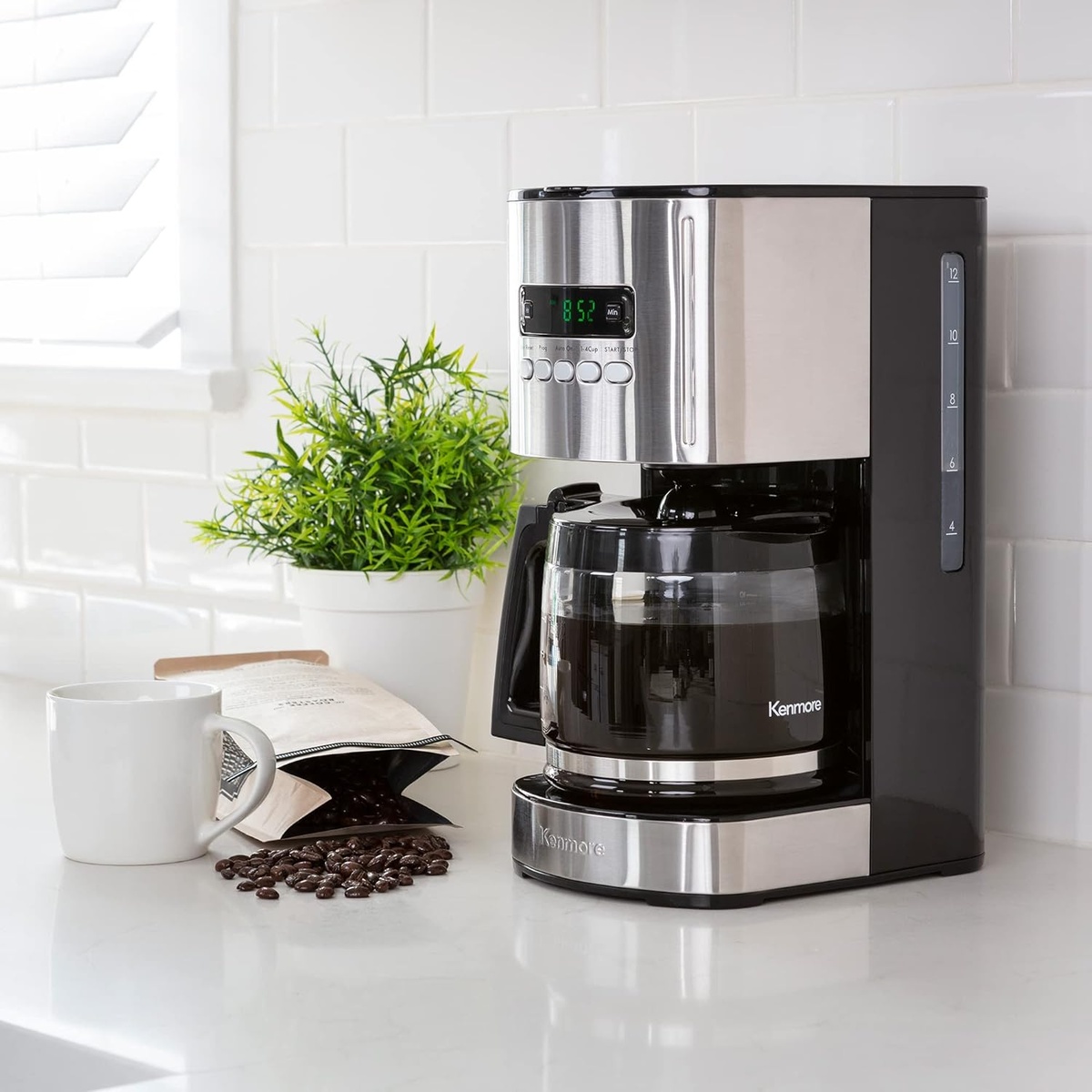 Kenmore drip coffee maker with glass carafe on a kitchen counter next to a mug and coffee beans