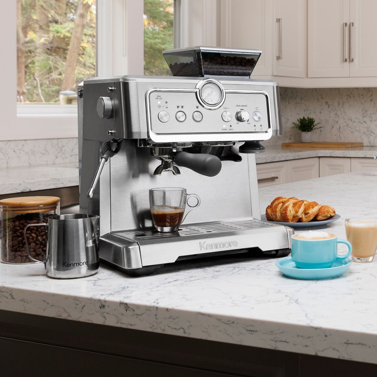Kenmore brushed stainless steel espresso machine on a kitchen countertop with espresso in a glass cup, a milk frothing pitcher, and prepared coffee drinks nearby.