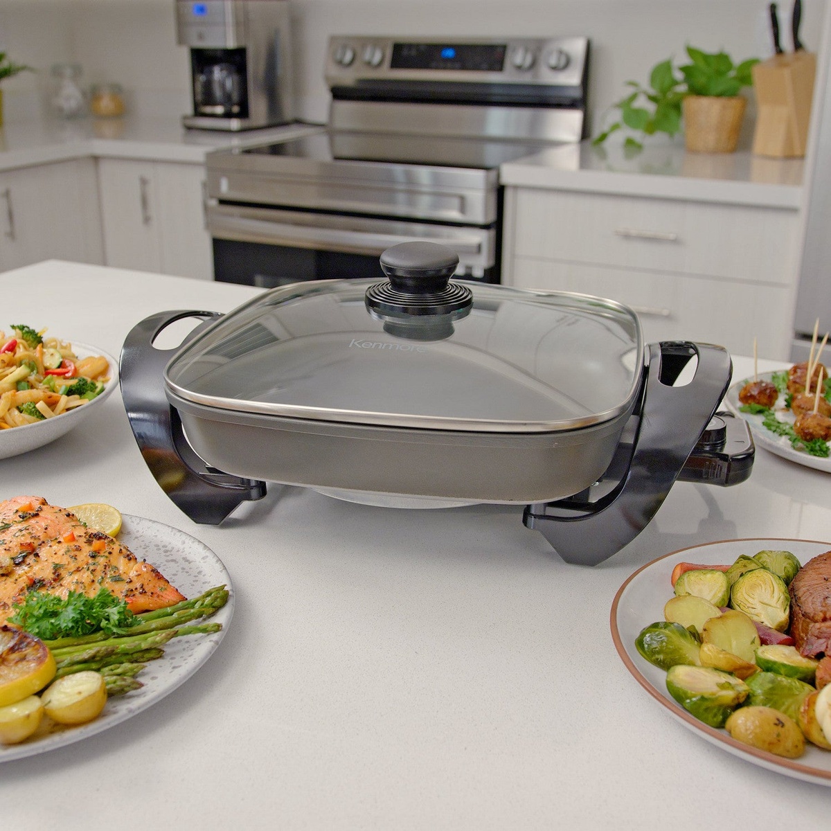 Kenmore electric skillet with glass lid on a kitchen countertop surrounded by plated food
