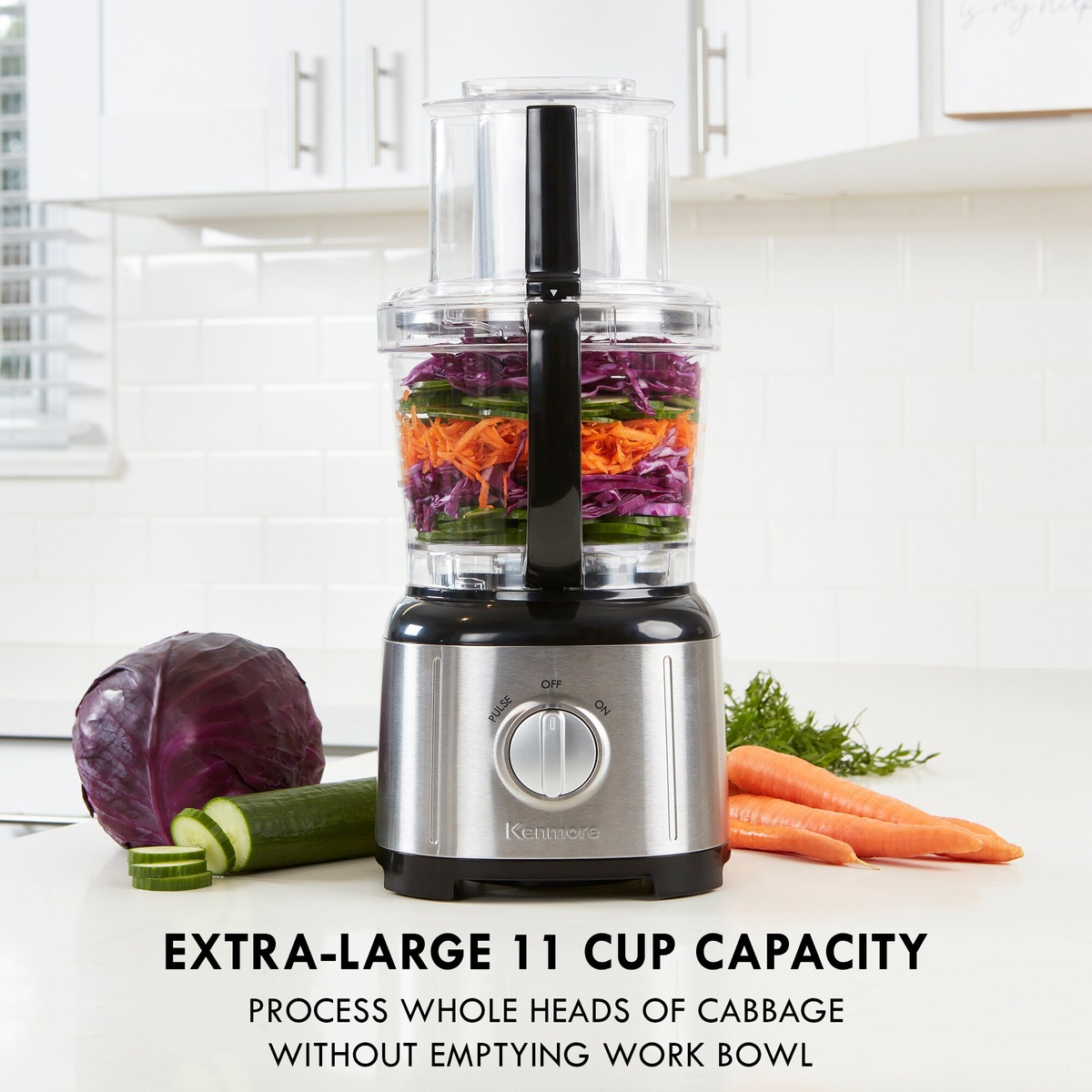 Kenmore food processor on a kitchen counter filled with shredded vegetables and surrounded by carrots, cucumber, and cabbage