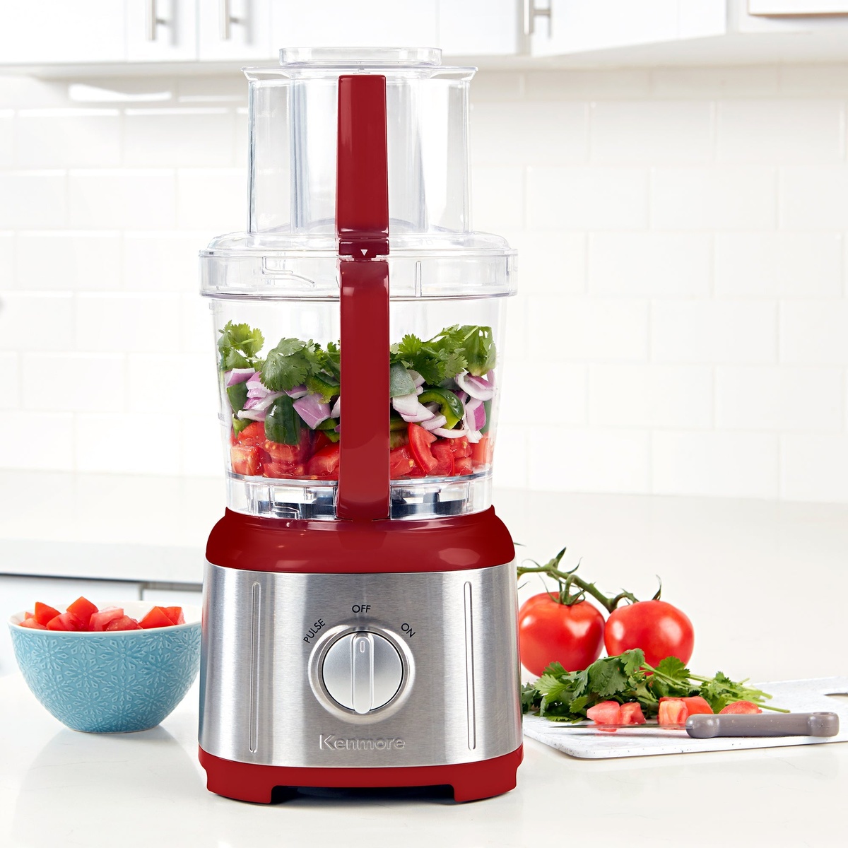 Kenmore red and stainless food processor filled with chopped ingredients on a bright kitchen counter with tomatoes and herbs