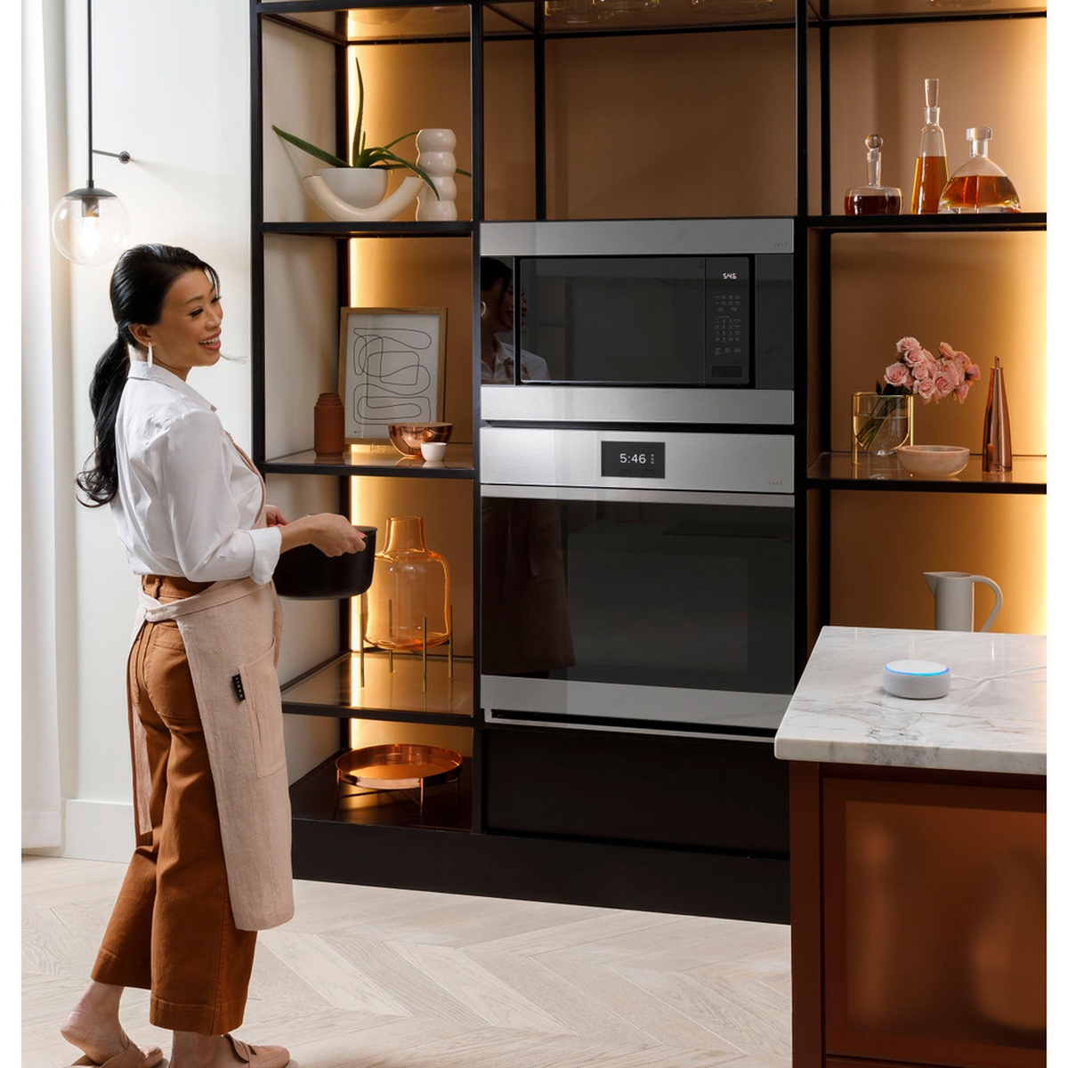 Lifestyle shot of a woman in a modern kitchen standing in front of a Café wall oven installed in a dark open-shelf unit with warm ambient lighting