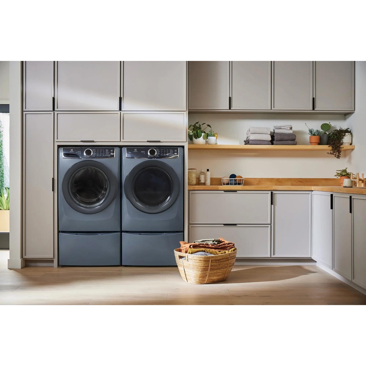 Electrolux denim blue washer and dryer pair installed on matching pedestals in a bright, modern laundry room with open shelving, cabinetry, and a wicker laundry basket in the foreground