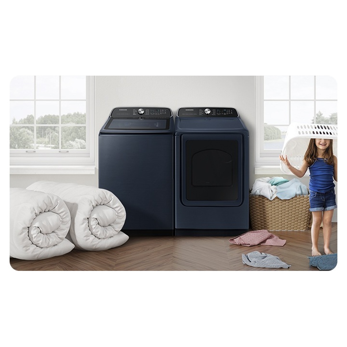 Samsung navy top load washer and matching dryer in a bright laundry room with rolled comforters in the foreground and a child carrying a laundry basket