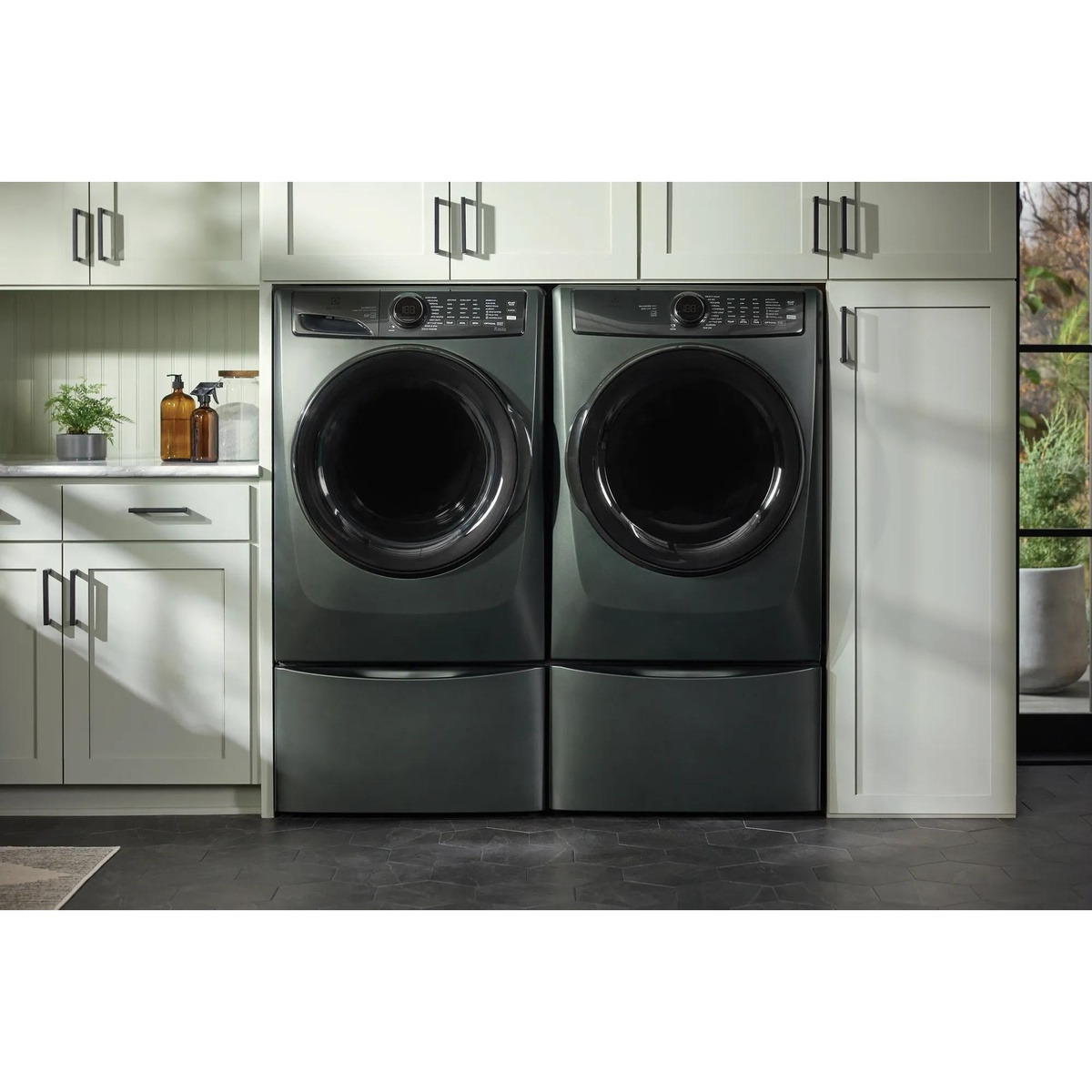 Electrolux front-load washer and dryer pair elevated on matching Alpine pedestals installed in a styled laundry room with white cabinetry