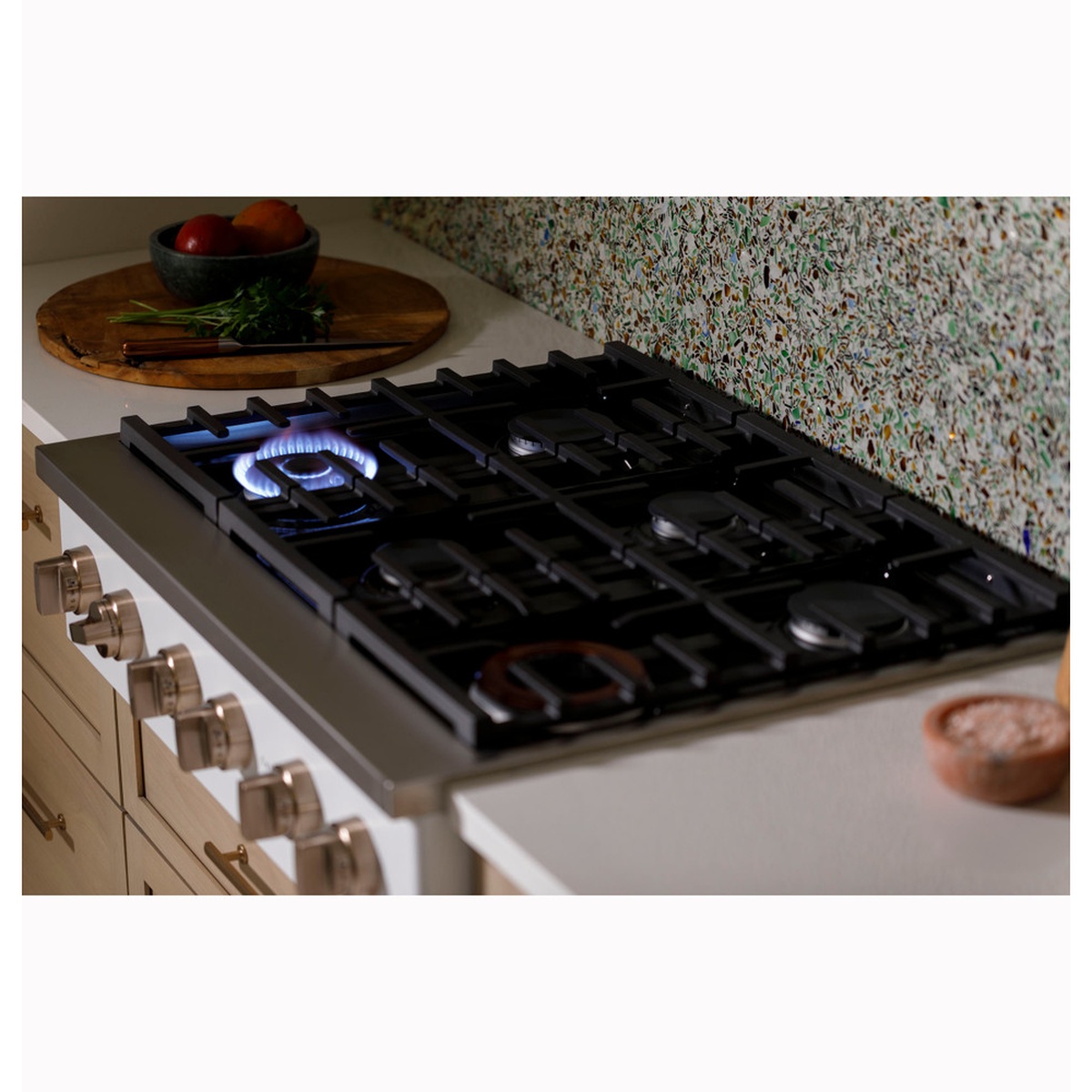 Lifestyle shot of Café rangetop installed in a modern kitchen with a blue gas flame lit on one burner, alongside a wooden cutting board with fresh produce