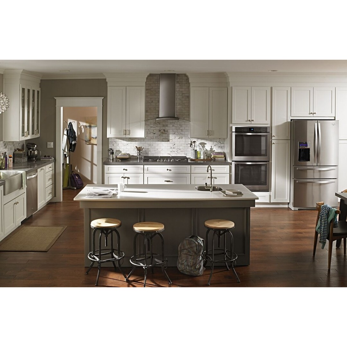 Lifestyle kitchen scene with wall-mount range hood installed above a gas cooktop, surrounded by white cabinetry, a kitchen island with bar stools, and stainless steel appliances