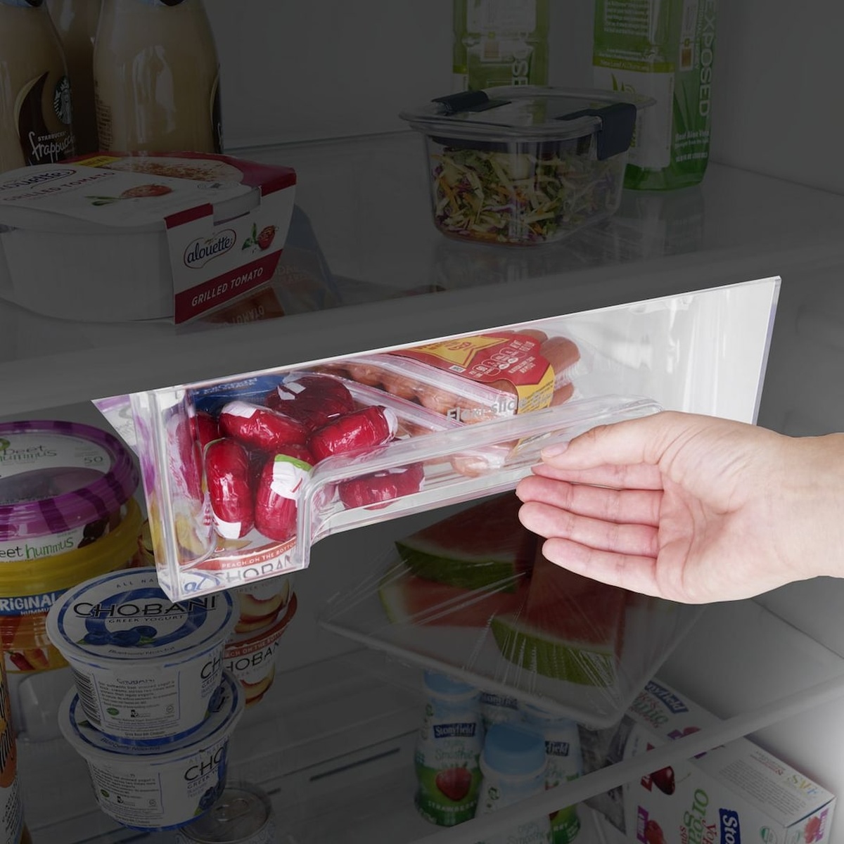 A hand sliding out the Flexi-Slide Deli Drawer inside the Whirlpool top freezer refrigerator, revealing packaged deli meats alongside shelves stocked with yogurt, hummus, and fresh food containers.