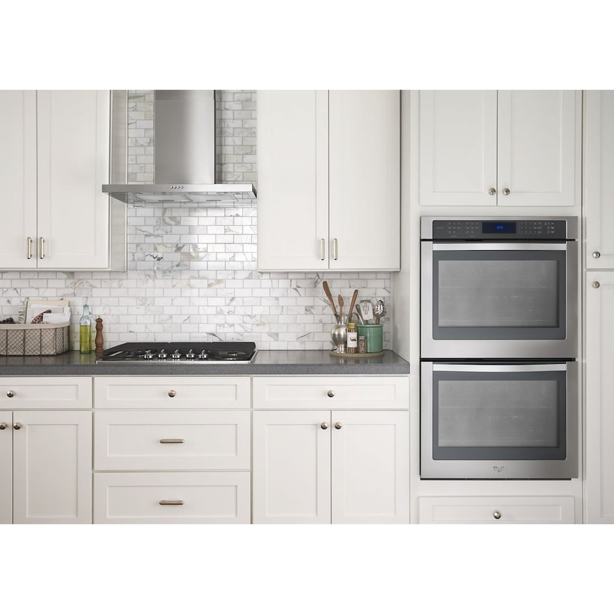 Whirlpool stainless steel wall mount range hood installed above a gas cooktop in a bright white kitchen with marble tile backsplash