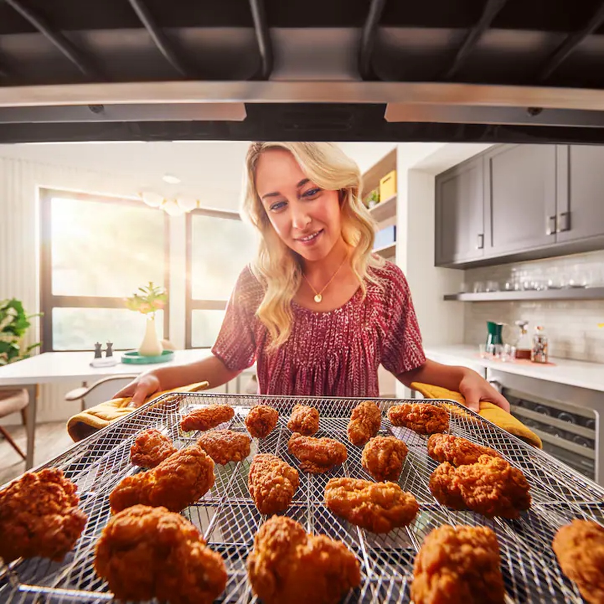 Woman using oven mitts to pull a wire basket filled with golden air-fried chicken pieces from a KitchenAid wall oven in a bright modern kitchen