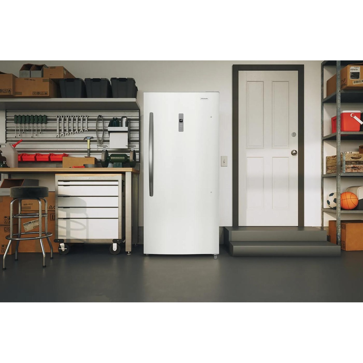 White Frigidaire upright freezer placed in a garage beside a workbench and storage shelves