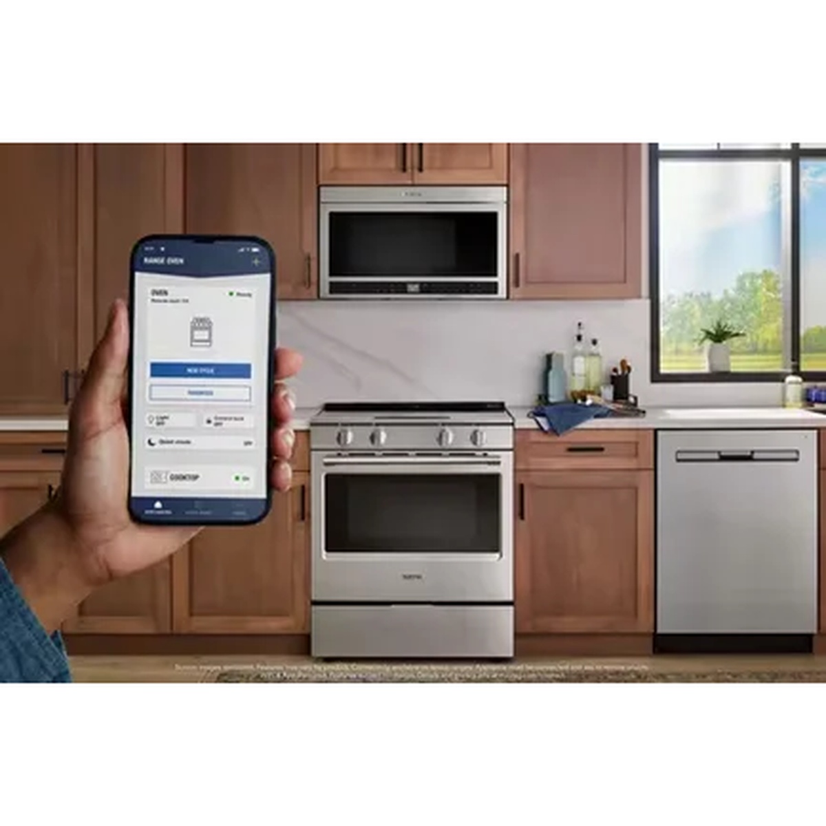 Hand holding a smartphone with the Maytag app in front of a stainless steel Maytag slide-in range installed in a kitchen