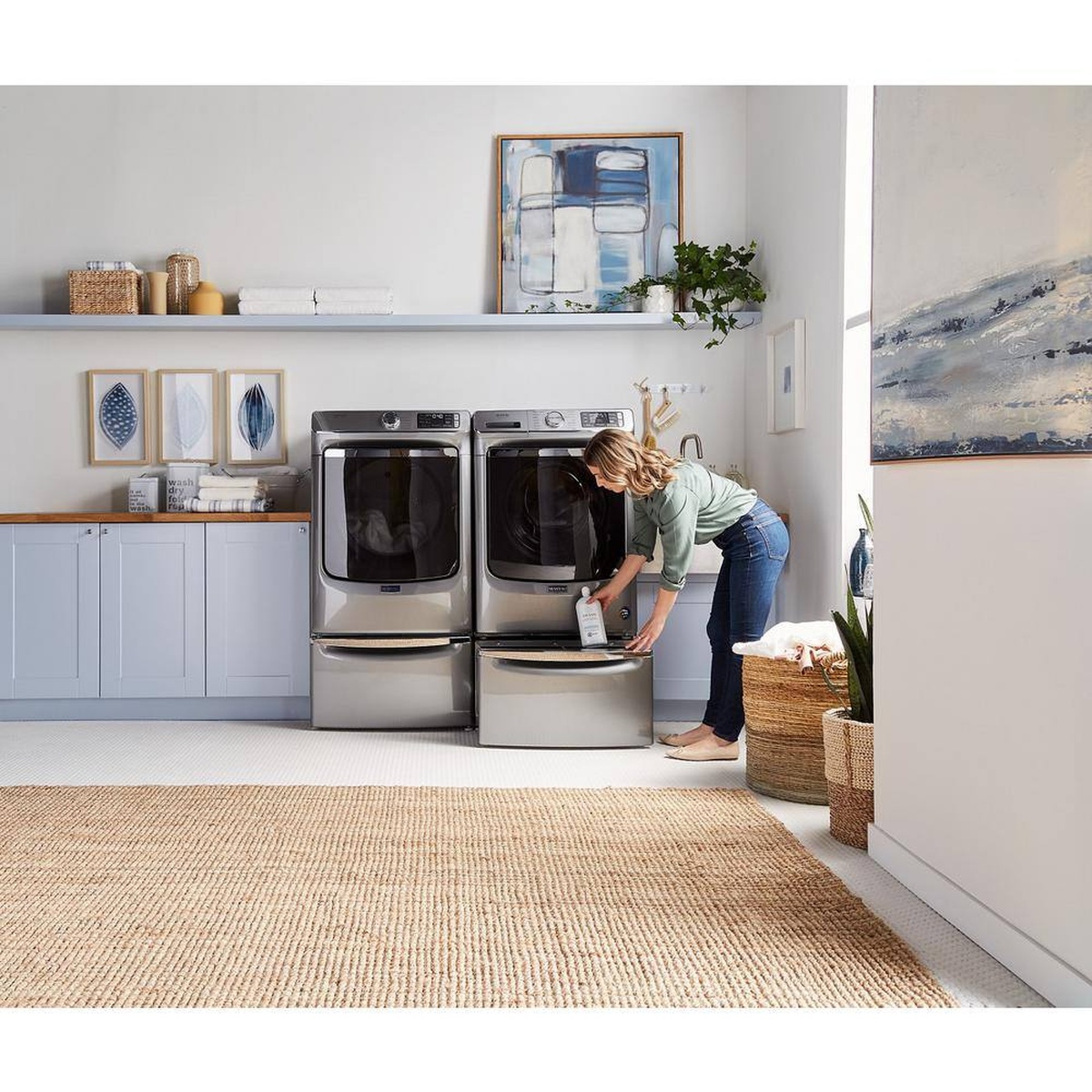 Laundry room scene with a front-load washer and dryer on matching pedestals; a person is reaching into the open pedestal drawer