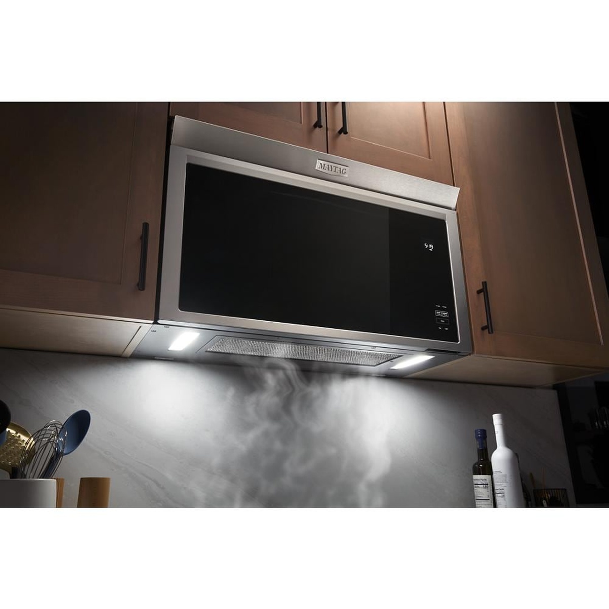 Maytag flush-mount over-the-range microwave installed under cabinets with underside lights on and the vent grille visible